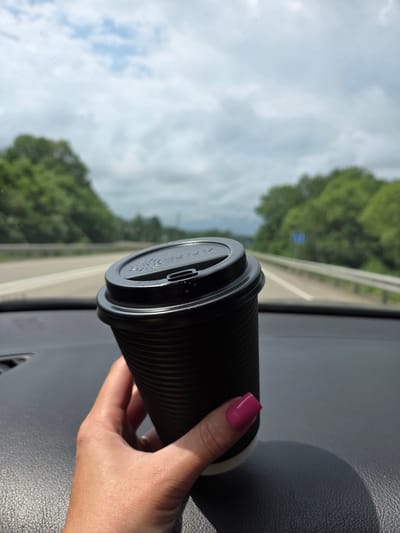 A hand with bright pink nail polish holds a black disposable coffee cup with a black lid. The cup is corrugated and appears to be made of paper. The hand and cup are positioned in the foreground, resting on a dark-colored dashboard. In the background, a highway stretches into the distance, flanked by lush green trees and a cloudy sky. There are no discernible buildings or specific landmarks of Obluchye, Russia, visible in the image.