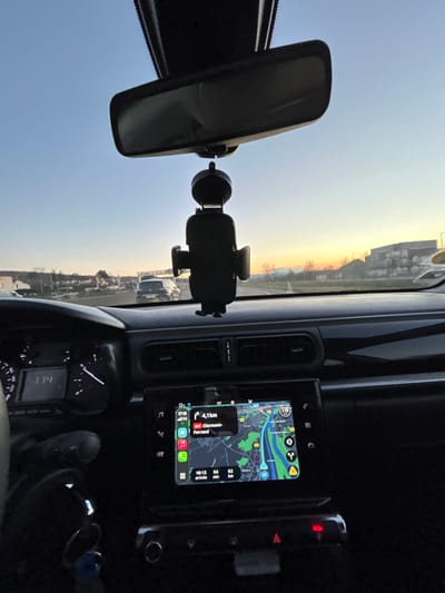 An interior view of a car, focusing on the dashboard and windshield. A rearview mirror hangs centrally, and a phone mount is attached to the windshield. A digital display shows a navigation map and driving information. A speedometer and other car controls are visible. The sky is visible through the windshield, with a gradient from blue to a warm sunset glow.