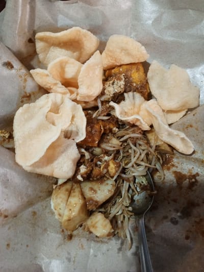 A close-up, overhead view shows a plate of Indonesian food, likely gado-gado, served on brown paper. The dish consists of vegetables, tofu, and crackers, all covered in a peanut sauce. A spoon is visible in the lower right corner of the image, partially submerged in the food. The food appears to be fresh and appetizing. The location in Ruwan Eliya, Sri Lanka is not discernible from the image itself.
