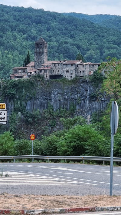 The image depicts the village of El Cós, Spain, situated atop a cliff overlooking a road. In the foreground, a road with white lane markings and a crosswalk is visible, bordered by a metal guardrail. A speed limit sign indicating "20" is positioned to the left of the road. The middle ground features a steep, rocky cliff face covered in green vegetation, with a cluster of stone buildings and a bell tower perched on the edge. The buildings have red-tiled roofs and stone walls. In the background, a dense forest covers a hillside. The sky is overcast.
