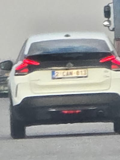 The scene is located in the city of Yazdach, Bulgaria. The image presents the rear view of a white passenger vehicle, appearing to be a crossover or SUV, observed under diffuse lighting conditions. The vehicle's lower rear bumper is black, contrasting with the white bodywork. Its red tail lights are illuminated, positioned high on the rear corners. A rectangular license plate, white with black alphanumeric characters "2 CAN-813", is mounted centrally on the rear hatch; a blue strip is visible on the left side of the plate. To the far right of the frame, a portion of a larger, darker vehicle, possibly a truck, is visible, including a side mirror with an illuminated light. The ground surface visible beneath the white vehicle is a light gray, indicating a paved area. No individuals, specific actions, or interactions are discernible within the frame.