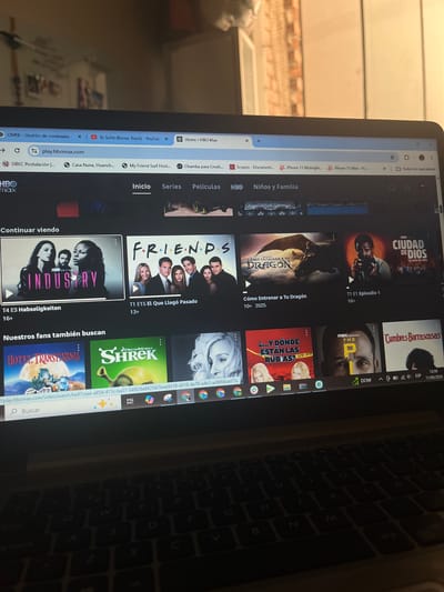The image shows a laptop screen displaying the HBO Max streaming service interface. The screen displays several movie and TV show posters, including "Industry," "Friends," and "How to Train Your Dragon." The user's cursor is hovering over the "Industry" poster. In the background, a dimly lit room is visible, with shelves containing various items. The overall scene suggests a user is browsing content on HBO Max. The image does not provide any information about the location being San Luis, Peru.