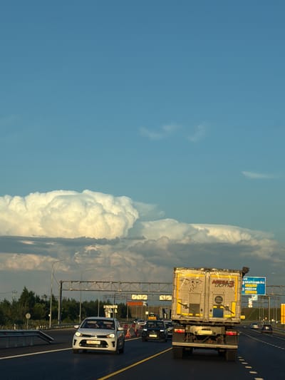 The image captures a sunny day in Egor'evo, Russia, with a clear blue sky dominating the upper portion and dramatic, large cumulonimbus clouds visible on the horizon. The scene is set on a multi-lane highway, with several vehicles in motion. In the foreground, a white sedan is traveling in the left lane, and a large white and yellow refrigerated truck is in the right lane. The truck has "MEGA LIGHT 3" written on its side and a drawing of a bull. In the background, other cars and trucks are visible, along with highway infrastructure including overhead signs and light poles. Trees line the sides of the highway. One blue highway sign in the distance indicates directions to Yekaterinburg, Moscow, and Tolyatti. The overall impression is one of a typical highway scene under a dynamic sky.