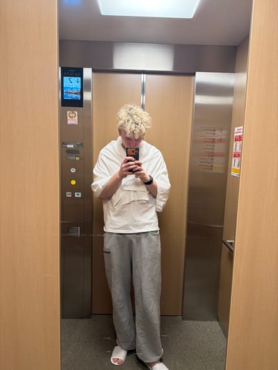 An individual with short, curly, blond hair is captured in a full-length mirror selfie inside an elevator, likely within the city of Kutchan, Japan, given the ski resort imagery on the elevator display. The person stands centrally, looking down at a dark-cased smartphone held in both hands, obscuring their face. They wear a white, textured, loose-fitting, short-sleeved garment, gray baggy sweatpants, and white open-toed slide sandals. A dark smartwatch is visible on their left wrist.

The elevator interior features light wood-paneled walls on the left and right, with a large mirror covering the wall opposite the observer. The floor is carpeted in a light, speckled gray. A recessed square light fixture is present in the ceiling. A stainless steel control panel is affixed to the left side of the mirror. It displays a digital interface showing a downward arrow, the number "1", and a graphic depicting a snowy mountain landscape with a ski lift. Below this, various buttons are present, one illuminated yellow, with all visible text primarily in Japanese. A second stainless steel panel on the right side of the mirror contains additional Japanese text and red-and-white informational graphics. A horizontal metal handrail is installed on the right wall, partially visible in the mirror. The elevator doors, visible as a reflection behind the person, are made of stainless steel.