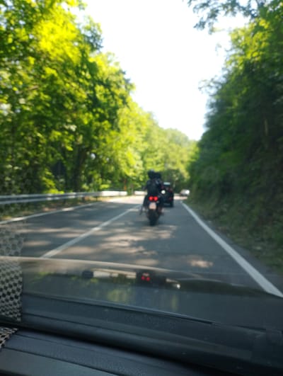 This is a slightly blurred, eye-level, long shot of a road with two motorcycles and a car traveling in the same direction. The road is paved and has white lines marking the lanes. It is flanked by lush green trees on both sides and a metal guardrail on the left. The sky is bright and appears to be a clear day.
The first motorcycle is in the foreground, and the rider is wearing a black helmet and a dark jacket. The second motorcycle is further ahead, and its rider is also wearing a black helmet. A dark-colored car is visible behind the second motorcycle. The scene appears to be on a scenic route, possibly in a rural or semi-rural area, given the abundance of trees.
While the image itself doesn't provide explicit clues to the location, the prompt specifies the city of Leonessa, Italy. Therefore, it can be inferred that this image was taken on a road in or near Leonessa, Italy.