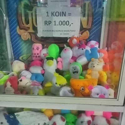 This image depicts the interior of a claw crane game machine, located in Kedung Dowo, Indonesia. The machine features a transparent glass front, behind which a dense collection of approximately 30-40 colorful plush toys is displayed. The toys are varied in size, color, and character, including recognizable figures such as Doraemon (green), Winnie the Pooh (yellow), a Spongebob-like character (orange), pigs (pink/white), ducks/penguins (white/yellow/grey), and other generic cartoon creatures in shades of pink, green, blue, yellow, white, orange, and red.
A white rectangular sign is adhered to the inside of the glass panel in the upper-center portion of the frame. The sign displays black text stating: "1 KOIN = RP 1.000,-" (indicating one coin costs 1,000 Indonesian Rupiah), followed by "SILAHKAN BELI KOIN DI KASIR/TOKO" (Please buy coins at the cashier/store), and "BY : TOYKIDS" at the bottom. The background inside the machine behind the toys features a colorful graphic design with diagonal stripes and patterned elements.
Below the main display compartment, a lower section or shelf is visible, also containing additional plush toys. The scene is brightly lit, consistent with an indoor arcade or retail environment. No people are visible.