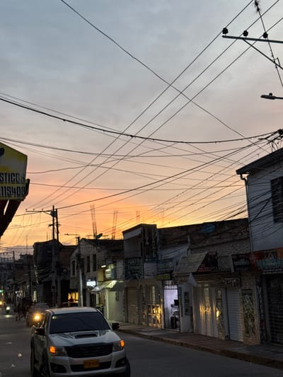 The image displays a street scene at dusk in Pitalito, Colombia. The sky transitions from a prominent orange-yellow hue at the horizon to a lighter blue-grey overhead. A dense, tangled network of black utility and power lines occupies the upper half of the frame, crisscrossing the sky.

In the foreground, slightly left of center, a light-colored pickup truck is positioned, facing the viewer. Its amber parking or turn signal lights are illuminated, and its license plate, RGN 458, is legible. The street surface is dark.

To the left, a partial yellow commercial sign with black text, displaying "STICO" and "115408," is visible. Below it stands a utility pole equipped with multiple cross-arms and insulators. Further down the street on the left, blurred figures indicative of pedestrians and additional vehicle lights are discernible.

The right side of the street is bordered by a row of two-story commercial buildings. Many of these storefronts are equipped with closed or partially closed metal security shutters. Several ground-level shops exhibit illuminated interiors, with one particular shop displaying what appears to be merchandise or a service counter. Visible signage on these buildings includes "PUNTO ELECTRONICS," "RADIOS," and "VARGAS." A blue satellite dish is mounted on one of the buildings. Rooflines vary, with some structures showing exposed rebar. A street light fixture extends from a pole near the upper right corner.