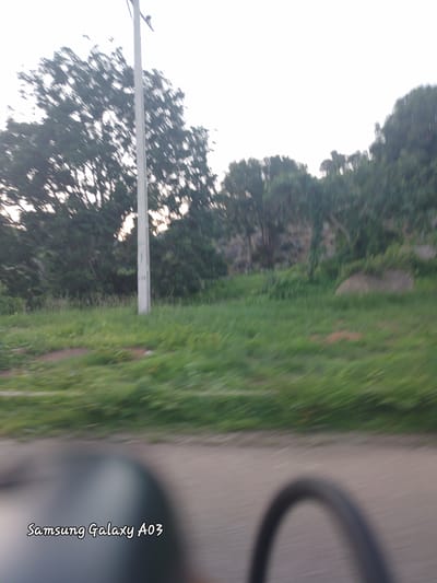 The image depicts a slightly blurry outdoor scene, likely captured while in motion. In the foreground, the bottom portion of the image shows a portion of a dark, curved object, possibly a bicycle wheel or part of a vehicle. The text "Samsung Galaxy A03" is superimposed in white cursive font in the lower left quadrant of the image.

The midground features a paved surface, and beyond that, a grassy area with sparse vegetation and a light-colored pole, possibly a utility pole or flagpole, standing vertically. In the background, a dense line of trees forms a natural backdrop, with a rocky, uneven terrain visible behind the trees on the right side of the frame. The sky is overcast and bright, suggesting it could be early morning or late afternoon. The overall impression is that of a roadside view with natural elements and some man-made structures. The provided context places this scene in the city of Gu, Nigeria.