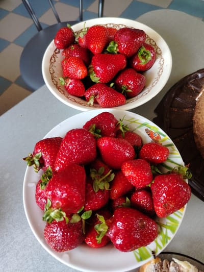 The image displays two white bowls filled with fresh strawberries, arranged on a light gray surface. A dark brown cake stand with a slice of cake is partially visible on the right side of the image. The background shows a patterned floor with blue and gray tiles and a dark chair. This scene is located in Rudzensk, Belarus.