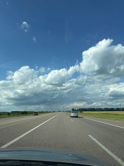 The image shows a view from a car traveling on a highway in Ksaverivka, Ukraine. The sky is filled with large, dramatic cumulus clouds against a bright blue background. The highway stretches into the distance, with a white dashed line dividing the lanes. Several cars are visible on the road ahead, including a silver car and a red car. The side of the road is lined with a field of green grass and a dense row of trees. The perspective suggests it is daytime, likely midday, given the bright sunlight illuminating the clouds and the road. The weather appears to be warm and partly cloudy. The scene conveys a sense of travel and the vastness of the open road under a dynamic sky.