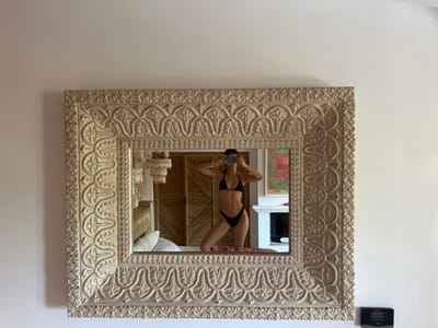 A woman wearing a black bikini is captured in a mirror's reflection, taking a selfie with her phone. She is standing on a bed, her body angled slightly towards the mirror. Her hair is dark and pulled back. Behind her, the room is visible, featuring a wooden headboard and a white pillow on the bed. To the right, a fireplace is visible, and above it, a framed artwork hangs on the wall. The mirror itself has an ornate, carved frame in a light, neutral color. A black electronic device is mounted on the white wall to the right of the mirror. The overall scene suggests an indoor setting, possibly a bedroom. The lighting is bright and appears to be natural, coming from an unseen source.