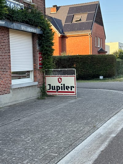 The image depicts a storefront with a red brick facade and a white rolling shutter. An awning with the text "STELLA ARTOIS" is visible above the shutter. To the right of the building, a white sign with a red border displays the word "Jupiler" in red lettering, accompanied by a lion emblem. A neatly trimmed hedge runs along the side of the building and extends behind the Jupiler sign. In the background, a red brick house with a dark-tiled roof covered in solar panels is visible. The scene is set on a paved walkway or sidewalk, with a road curving to the right in the foreground. The lighting suggests it is daytime, with sunlight casting shadows. This image was taken in Tessenderlo, Belgium.