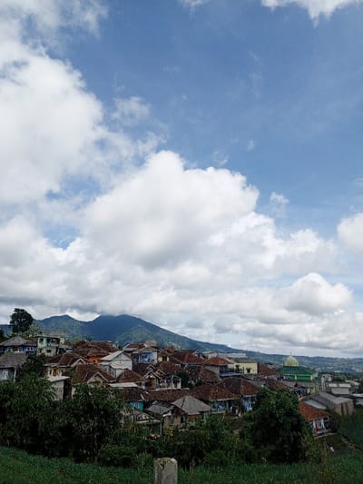 A mountain rises behind a village of houses with red tile roofs, interspersed with greenery. A green-domed mosque is visible. The sky is a vibrant blue, filled with large, puffy white clouds that dominate the upper portion of the frame. The overall impression is one of a peaceful, rural landscape under a dynamic sky.