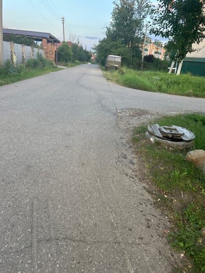 The image depicts a residential street in Apsheronsk, Russia, likely during the daytime due to the ambient light. The street is paved with asphalt and shows signs of wear, including cracks and patching. To the left, a fence made of stone and metal panels borders a property with lush green vegetation and a tall white flower. A telephone pole with multiple wires is visible behind the fence. Further down the street, a similar fence encloses more greenery.
On the right side of the road, overgrown grass lines the shoulder. A concrete manhole cover, partially broken and made of weathered wood and metal, is situated near the edge of the asphalt, surrounded by grass and a large stone. Further along this side, a small, utilitarian grey van is parked. In the background, a multi-story building with white and orange facades and green accents is visible, surrounded by trees. Another, taller building with a green roof and white brickwork stands to its right, with a white ladder leaning against it. The sky above is a clear, pale blue, suggesting fair weather.