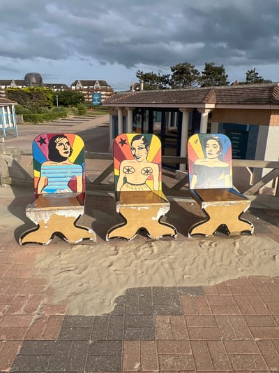 Three colorful, custom-designed chairs are displayed outdoors on a sandy area next to a brick pathway. Each chair features a painted portrait of a woman. The chair on the left depicts a woman with dark hair and a blue and striped top. The middle chair shows a woman with dark hair and a bandeau top. The chair on the right shows a woman with dark hair and an off-the-shoulder black top. The chairs are in front of a building with a tiled roof and a wooden fence. In the background, buildings and trees are visible under a cloudy sky. A large, abstract sculpture of a head is also visible in the distance, to the left of the frame. The scene appears to be located in Deauville, France, near a beach or promenade.