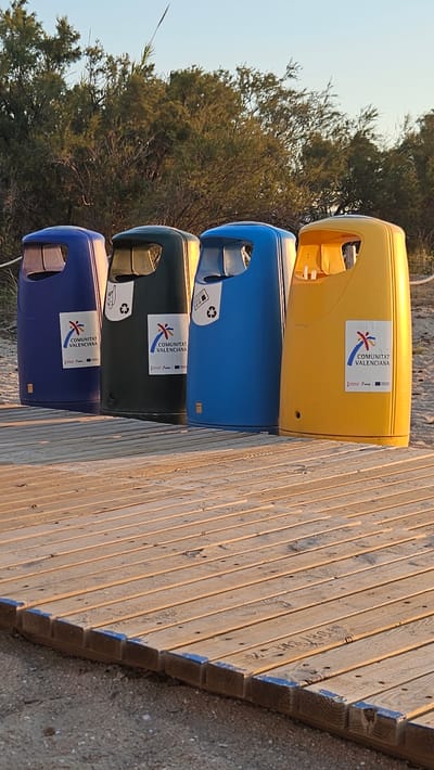 This media file shows a row of four colorful recycling bins on a wooden boardwalk. The bins are stacked vertically, with a yellow bin on top, followed by blue, dark green, and another blue bin at the bottom. Each bin has an opening for waste disposal. Some of the bins have stickers with a logo that reads "COMUNITAT VALENCIANA" and some recycling symbols.
The scene is set outdoors, with trees and vegetation in the background. The ground appears to be sandy, consistent with a beach or park setting. The wooden boardwalk is made of planks and runs diagonally through the frame, suggesting it might lead to a beach or other area.
The lighting suggests it is either early morning or late afternoon, with soft sunlight casting shadows and highlighting the texture of the wood. The sky is clear and pale, indicating fair weather. There are no people visible in the image. The overall impression is one of an organized public space, likely a beach area given the location context of Deveses, Spain. The bins are prominently displayed objects, representing a focus on waste management and environmental consciousness in the setting.