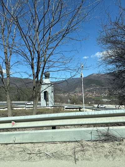The image depicts an outdoor scene in Bunovo, Bulgaria, under a clear blue sky, indicative of late winter or early spring. In the immediate foreground, a three-tiered metallic gray guardrail is present, obscuring the ground level, which consists of dry, light brown earth with sparse, dead vegetation.

Dominating the midground is a light gray stone monument. It features a tall, rectangular base with an arched opening, topped by a dark, rounded bust-like sculpture. This monument is positioned behind the guardrail and partially framed by several tall, bare deciduous trees with visible branches and twigs. To the right of the monument, a highway with multiple lanes is visible, on which a white car or SUV is traveling leftward. Further right, another white vehicle is also on the highway. A slender, white pole equipped with several antennae extends vertically into the sky, situated to the right of the monument.

The background reveals a wide valley landscape. A town, identified as Bunovo, is situated in the valley to the right, characterized by numerous buildings with visible roofs. Behind the town and monument, rolling hills and mountains, primarily covered in bare or sparsely vegetated trees, rise towards the horizon. A prominent concrete viaduct or bridge, featuring multiple arches, spans a valley in the mid-distance to the left of the town. The sky is bright blue with a few scattered, small white clouds. No individuals are visible in the scene.
