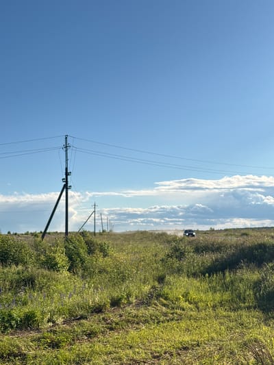 This is a vertically oriented, full-shot photograph taken outdoors in what appears to be a rural or natural setting in Ostrovskoye, Russia. The scene is dominated by a bright blue sky with scattered white, puffy clouds. In the foreground, a dark, tall utility pole with multiple wires stretches upwards. Behind the pole, to the right, a dense green field of tall grass and bushes lines a dirt road or path. Further in the distance, more utility poles are visible marching along the horizon, with a dark-colored vehicle, possibly a car, driving on the path. The light suggests it is daytime, likely mid-morning or afternoon, with clear and sunny weather. The overall mood is peaceful and open, characteristic of a rural landscape on a pleasant day. There is no visible text in the image.