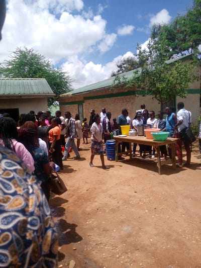 This outdoor image captures a bustling community gathering in Mwingi, Kenya, under a bright, partly cloudy sky. The scene unfolds on a reddish-brown dirt ground, characteristic of many local settings.
Numerous individuals, mostly adults, are congregated across the frame. On the left side, a dense crowd appears to be forming a queue or waiting, partially obscured by a person in the immediate foreground wearing a patterned blue and brown garment. In the center-right, a smaller group of people is actively gathered around a rustic wooden table.
The table serves as a central point, holding several items indicative of a serving or distribution event. These include large silver thermal containers or dispensers, a bright yellow plastic bucket, and distinct green and pink plastic basins. A blue plastic barrel or drum is positioned on the ground next to the table. People around the table are interacting, some appearing to be serving or receiving items.
In the background, sturdy buildings with light-colored walls – one with a horizontal green stripe – and green corrugated metal roofs are visible. Lush green trees flank and tower over the structures, adding to the natural environment.
The bright sunlight, casting clear shadows on the ground, suggests it is midday or early afternoon, with comfortable weather conditions. The overall atmosphere appears orderly and engaged, with people participating in what seems to be a communal meal or provision distribution. No discernible text is visible on any objects or clothing within the image.
