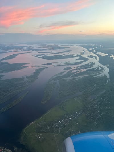 An aerial view shows a wide, winding river system with numerous islands covered in dense green trees. The river's surface reflects the soft pastel colors of a sky with pink and pale blue clouds. On the right bank, a small village with scattered houses and green fields is visible. A portion of an airplane's blue and white engine is in the bottom right corner of the frame. The scene appears to be captured during twilight. The geographic location is the city of Kokkorevo, Russia, where the Keret River flows.