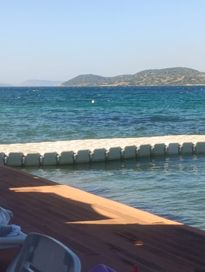 The image captures a serene coastal scene at a location that appears to be Urla, Turkey, specifically a waterfront area likely featuring a pier or deck. The foreground is dominated by a wooden deck, partially in shadow, with some white beach chairs visible on the left. Beyond the deck, a modular floating platform system, composed of interconnected white plastic cubes, extends into the turquoise water, serving as a walkway or extension of the shoreline. The sea is calm with gentle ripples, and in the distance, rolling hills with sparse vegetation form the coastline. The sky is clear and blue, indicating fair weather. The overall impression is one of a tranquil seaside leisure spot.