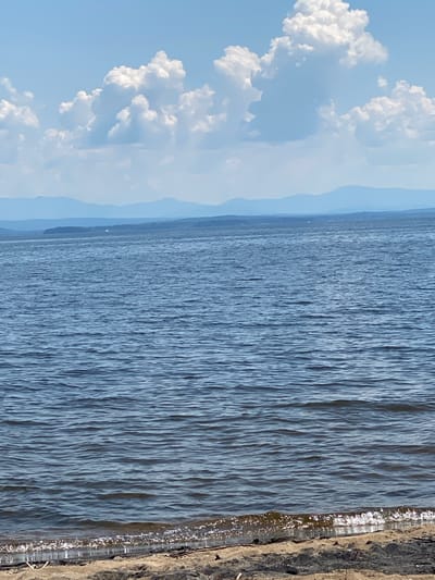 The image depicts a serene natural scene of a body of water, likely a lake or large river, with a distant shoreline and a bright sky with fluffy white clouds. The water is a deep blue and shows gentle ripples, suggesting a calm day. On the right side of the image, a textured embankment or shoreline is visible, with some dry vegetation or debris. The sky occupies the left portion of the image and is a light blue, adorned with large, puffy clouds that appear bright and illuminated, indicative of daylight. Faint, hazy mountains or landforms can be seen in the distance, blurring into the horizon. The perspective is from a slightly elevated position looking out over the water. There is no text visible in the image, and no people or specific objects are discernible. The overall mood is peaceful and tranquil, suggesting a pleasant day outdoors in a natural setting, possibly in the Town of Peru, USA. The time of day appears to be midday or afternoon, given the bright illumination and the appearance of the clouds.