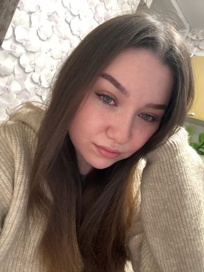 A young woman with long, dark brown hair and green eyes is shown in a close-up, head-and-shoulders shot. She is wearing a beige knitted sweater and leaning her head on her arm. The background is a wall with a floral wallpaper pattern in shades of white and grey. The lighting suggests an indoor setting. There is no discernible location in the city of Sakmary, Russia.