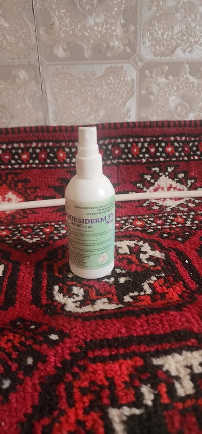 An overhead shot shows a white spray bottle labeled "ROKSIDERM 3%" resting on a red and black patterned rug. The rug is placed on a floor with light-colored, textured wallpaper. The bottle, which is partially obscured by the patterned rug, has text in what appears to be Cyrillic and Latin alphabets. The lighting is diffused, suggesting an indoor setting. The overall scene is still and lacks any visible human presence or activity. The location in the city of Türkmenbaşy, Turkmenistan, is not discernible from the provided image.