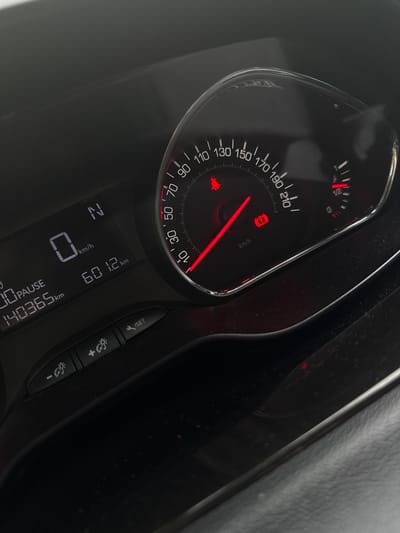 The image displays a close-up view of a car's dashboard. Prominently featured is the speedometer, which shows a needle pointing to 0 km/h. To the left of the speedometer is a digital display indicating "N" for neutral gear, a speed of 0 km/h, and an odometer reading of 140365 km. Below this, it shows "PAUSE" and "601.2 km". The dashboard also has several indicator lights illuminated in red, including what appears to be an airbag warning light and a brake system warning light. Below the main displays are several buttons with symbols indicating light adjustment and a "SET" function. The environment appears to be an interior shot of a car, suggesting the vehicle is stationary. There is no indication of the city of Amstelveen, Netherlands, in the image.