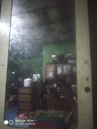 The image depicts a distorted reflection captured in a dusty or smudged mirror, providing a view of an interior room within a location in Lumajang, Indonesia. The reflected scene shows a room with light green painted walls. On the left side, a light-brown wooden chest of drawers with at least three visible drawers is present. On its surface rests a cylindrical pink and white patterned container and a white, possibly an alarm clock or decorative item. To the right of this, a wooden display cabinet with glass doors is visible, holding several framed pictures or objects. The floor is covered with a patterned textile, possibly a rug, displaying blue, red, and darker tones, with indistinct objects scattered upon it. A human arm and hand, holding a smartphone with the visible watermark "SHOT ON REDMI 7 AI DUAL CAMERA," are observed in the lower right quadrant, taking the photograph. The mirror itself is bordered by a light-colored, vertical frame on the left and right edges.