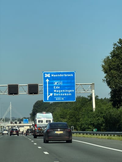 The image shows a Dutch highway with a blue directional sign indicating "Maanderbroek" and "N24" with destinations "Ede", "Wageningen", and "Bennekom" 600 meters ahead. The sign also shows a lane split, with the left lane going straight and the right lane turning. Multiple vehicles are on the highway, including a black Tesla, a white van, and several other cars. The environment is sunny with a clear blue sky and trees lining the side of the road. The sign is positioned over the highway, with traffic lights on either side. The image appears to have been taken from a moving vehicle, as indicated by the slight motion blur. A green sign with "N233 110.3" is visible on the right side of the highway. The overall scene depicts a typical highway environment in the Netherlands, with clear signage and moderate traffic.