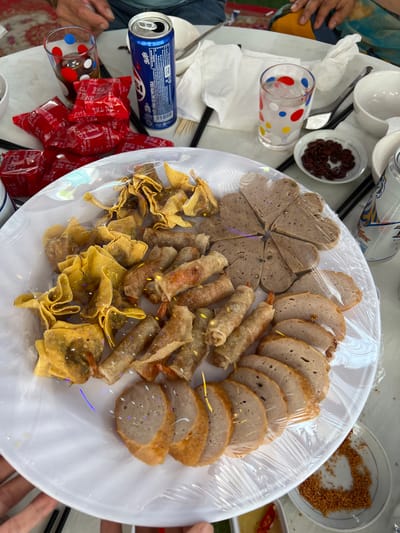 A high-angle shot captures a casual meal spread on a light-colored table, likely in Bình An, Vietnam. The central focus is a large white platter, covered with transparent plastic wrap, laden with various Vietnamese dishes. These include heart-shaped and round slices of a light brown, sausage-like item (possibly chả lụa or giò lụa), fried spring rolls, and crispy fried wonton-like snacks.

Around the platter, several hands are visible, suggesting people are gathering for the meal. Beverages include an open blue Pepsi-Cola can and a silver Tiger Beer can, along with a clear glass with colorful polka dots holding a dark drink. Tableware consists of white bowls, small plates (one containing dark reddish-brown seeds, another with what appears to be toasted coconut or peanut flakes), chopsticks, and white napkins. A small dish holds dipping sauce with sliced red chilies.

Next to the Pepsi can are several small red packets, likely containing condiments or snacks, with Vietnamese text such as "CÒN NHỚ THU CHO CẤP" and "AN THO". The Pepsi can itself has "NƯỚC UỐNG CÓ GAZ PEPSI COLA" printed on it. The bright lighting suggests it is daytime. The entire scene depicts a convivial gathering enjoying a selection of local Vietnamese food.