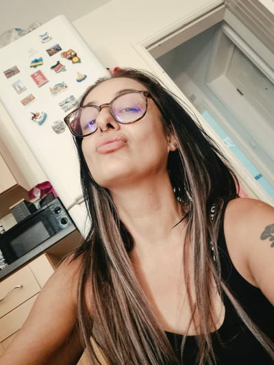 A close-up, high-angle, selfie of a young woman with long, dark, streaked hair, wearing glasses and a nose ring, is taken in what appears to be a kitchen. The woman is puckering her lips and looking upwards. Behind her is a white refrigerator covered in various colorful magnets, some of which appear to be travel-related. To the left of the refrigerator is a microwave oven, and beyond that, kitchen cabinets. To the right of the woman is a slightly ajar white door, possibly to a closet or another room, with a light fixture visible above it. The image is lit by natural light coming from an unseen source. The overall setting suggests an indoor domestic environment. There is no specific indication of the location being in Bridlington, United Kingdom.