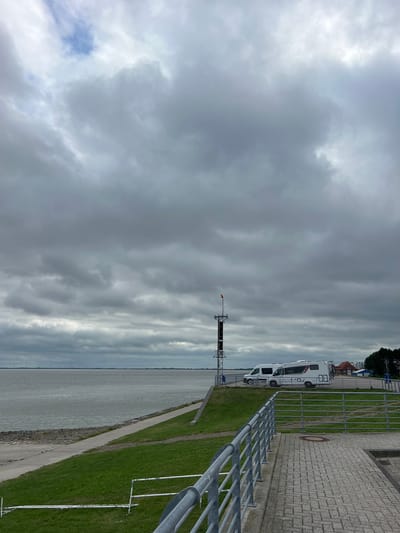 The image captures a cloudy day at the Jade Bay in Wilhelmshaven, Germany. In the foreground, a paved walkway with a metal railing runs diagonally across the frame. To the left, a grassy embankment slopes down towards the water. A concrete path is visible on the embankment. The water is calm and stretches to the horizon, where a line of distant wind turbines can be seen. On the raised area behind the embankment, two white camper vans are parked. A tall communication tower with a small orange flag at its top stands near the camper vans. In the background, buildings and trees are visible on the far shore.