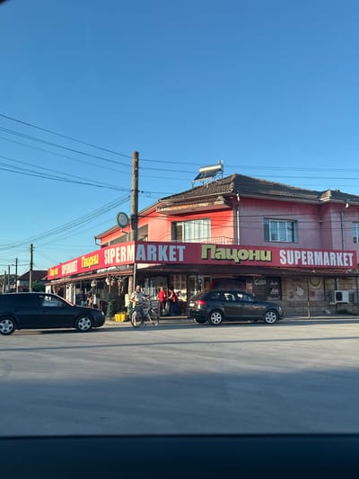 The image captures a street scene in the city of Tsar Kaloyan, Bulgaria, featuring a prominent red building with a large banner that reads "SUPERMARKET". The banner also includes Cyrillic text, "Пацони". The building appears to be a commercial establishment, likely a grocery store, with visible windows and an air conditioning unit mounted on the exterior wall. In front of the supermarket, there are several people. One individual is on a bicycle, and two others are standing near the entrance. Two cars are parked or driving on the street in front of the building; one black car is on the left, and a dark grey hatchback is on the right. The sky is clear and blue, suggesting a sunny day. Electrical wires are visible suspended across the sky, supported by utility poles. The foreground shows a paved road or parking area.