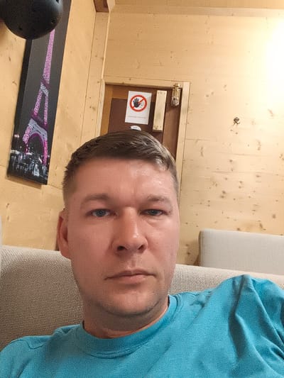 The media file is a selfie taken by a man sitting on a couch. He is wearing a blue t-shirt and has short brown hair. The background consists of wooden walls, a framed picture of the Eiffel Tower, a door with a "no entry" sign, and part of a light-colored couch. The lighting appears to be artificial, suggesting the photo was taken indoors. The overall mood is casual and relaxed. There is text on the "no entry" sign, but it is too small to read clearly. The provided location context suggests the photo was taken in Wädenswil, Switzerland.
