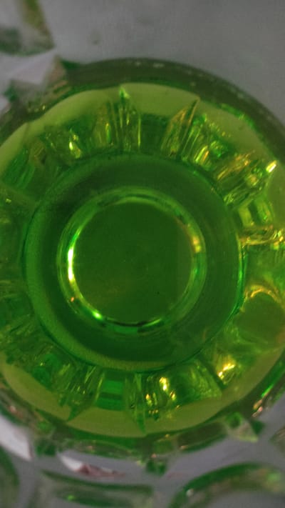 This is a close-up, top-down view of a green glass. The glass appears to be transparent with a textured rim. The lighting creates reflections and highlights on the surface of the glass, giving it a glossy appearance. There is no visible indication of location within the city of Bolotnoye, Russia. The image solely focuses on the glass object.