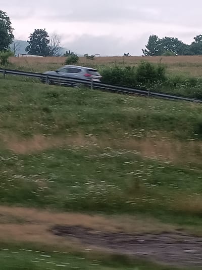 A gray SUV is driving on a paved road in Brețcu, Romania. The SUV is on the right side of the road. The road is bordered by a grassy area with some white flowers. In the background, there are trees and hills. The sky is cloudy.