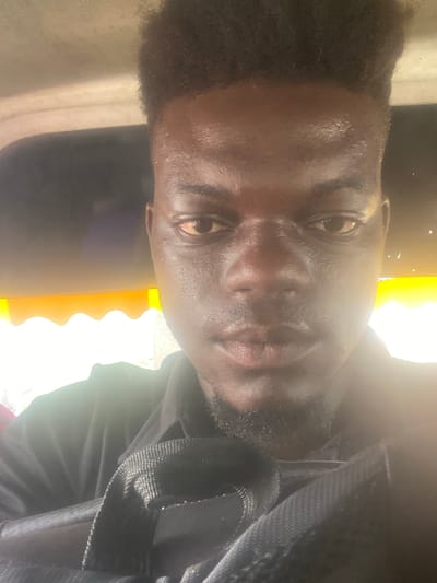 The image is a portrait of a dark-skinned man, rotated 90 degrees counter-clockwise, showing him looking directly at the camera. He has short, dark, kinky hair and a short beard. His face appears slightly shiny, possibly due to sweat or natural oils, particularly on his forehead and the bridge of his nose.
He is wearing a dark-colored garment, likely a black shirt or jacket. A black, woven fabric strap, possibly from a backpack or bag, is visible across his right shoulder (when the image is correctly oriented).
The background is composed of contrasting light and dark areas. A very bright, yellow-orange light source is prominent on the left side of the frame (bottom if rotated correctly), which creates a strong glow and causes some areas to be overexposed. This bright section has a distinctive wavy or scalloped edge. The right side of the background (top if rotated correctly) is mostly dark.
The strong light suggests it is daytime. The man has a neutral to slightly serious expression. He appears to be posing for a photograph or taking a selfie. There is no visible text in the image.