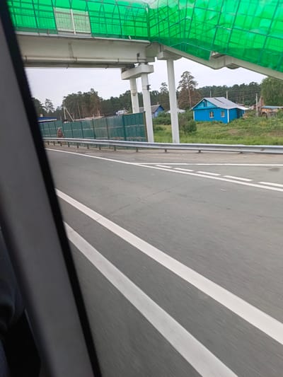 A view from a vehicle traveling on a road shows a green pedestrian overpass with a concrete structure supporting it overhead. The road surface is asphalt with white lane markings. To the right of the road, a turquoise metal fence runs along the side, backed by green grass and trees. A small blue house with a gray roof is visible amongst the greenery. Further in the distance, more trees form a treeline. The sky is overcast.