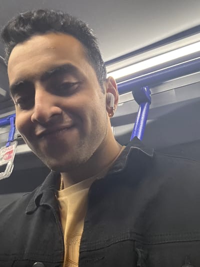 A smiling man with short, dark hair wears a black denim jacket over a tan or light gold-colored shirt. He has an earbud in his left ear. Behind him are bright lights and a blue handrail. The background is out of focus.