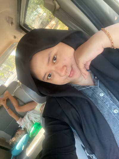 This is a selfie of a person wearing a black hijab and a denim shirt, taken inside a vehicle. The person's right hand rests on their chin, and they are smiling. In the background, a child is lying down in the back seat, appearing to be asleep or resting, with two colorful pillows next to them. The interior of the car is visible, including the seats and a sunroof. Through the sunroof and windows, trees and sunlight suggest an outdoor environment. The image appears to have been taken during the daytime. The location is identified as Ngrambe, Indonesia.