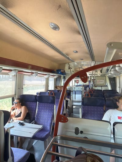 The image shows the interior of a train carriage with rows of empty and occupied seats. The seats are upholstered in a purple fabric with a patterned design. In the foreground on the left, a woman wearing sunglasses and a black top is seated at a table, looking out the window. She has her chin resting on her hand. To the right of the woman, another seat is occupied by a person wearing a white t-shirt. The train carriage has overhead luggage racks and visible lighting fixtures. The windows reveal a blurred view of greenery outside, suggesting the train is in motion. The color palette of the interior is dominated by beige on the ceiling and the purple of the seats, with touches of red on the overhead handles. The scene is generic to train travel and does not contain any specific visual cues to pinpoint its location in Elne, France.