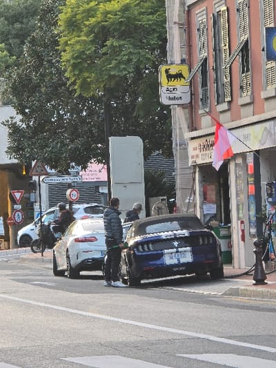 A street scene shows a blue Ford Mustang parked next to a white BMW. People are standing near the cars. A flagpole with a white and red flag is on the building. A gas station sign and several traffic signs are visible on the left side of the frame. A tree with green leaves overhangs the street. The image is slightly rotated, resulting in a distorted perspective.