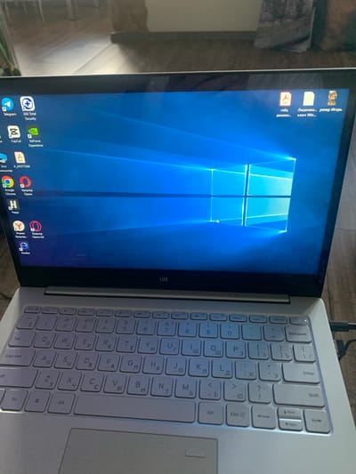 The image shows a laptop with a blue Windows wallpaper. Several icons are visible on the desktop, including Telegram, 360 Total Security, CapCut, GeForce Experience, a PDF file, and others. The keyboard is partially visible, and some of the keys have Cyrillic characters on them. A power cable is plugged into the side of the laptop. The background suggests an indoor environment, possibly a room. There is no indication of the city of Buzlanovo, Russia, in the image.
