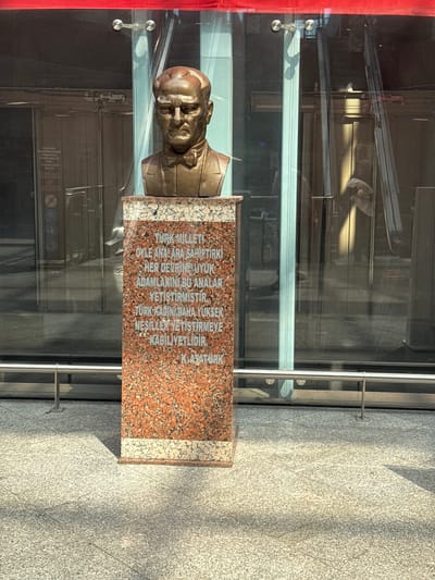 The image shows a bronze bust of a man, presumably a historical figure, mounted on a marble pedestal. The bust depicts a stern-faced man wearing a suit and bow tie. The pedestal has an inscription in Turkish. Behind the bust, a modern glass facade of a building is visible, with doors and reflections of the surroundings. The lighting suggests it is daytime, with shadows cast on the ground. The inscription on the pedestal translates to: "The Turkish nation has mothers who have raised great men of every era. The Turkish woman is capable of raising higher generations." The lower part of the inscription reads "K. Atatürk," identifying the bust as Mustafa Kemal Atatürk, the founder of the Republic of Turkey. The location is specified as Çankaya, Turkey.