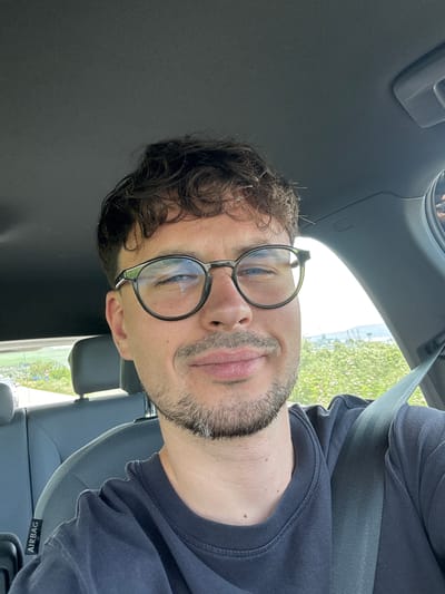 A close-up photo depicts a man with brown curly hair and a light beard wearing round-rimmed glasses. He is sitting in the driver's seat of a car, with the dashboard and seat visible. He is wearing a dark blue shirt and a seatbelt. The car's interior is predominantly gray. The word "AIRBAG" is visible on a tag near the bottom right of the frame. The scene is inside the car, with a view of foliage through the window. The exact location in the city of Ezerovo, Bulgaria, cannot be determined from the image.