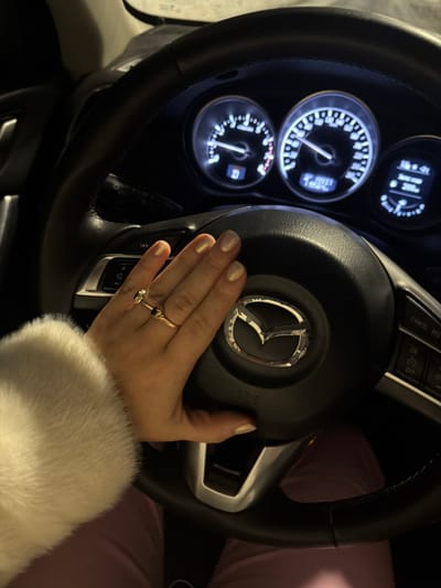A right hand, adorned with two gold-colored rings on the ring finger (one a simple band, the other featuring a dark central stone), is pressing the central horn pad of a black Mazda steering wheel. The fingernails exhibit a light, pearlescent polish. The driver's right forearm is partially visible, covered by a white, fluffy material, consistent with a fur-lined garment. Below the steering wheel, a segment of the driver's leg or lap, clad in light pink or purple fabric, is discernible. The illuminated dashboard, visible beyond the steering wheel, displays a speedometer indicating approximately 40 km/h and a tachometer. A digital readout shows an odometer reading of 99337, a trip meter of 155 km, and an external temperature of -15°C. The gear indicator 'D' is illuminated. The scene depicts the interior of a vehicle operating at night or in low-light conditions. The specific location within Азьмушкино, Russia, cannot be determined from the provided image.