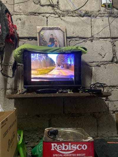 The image depicts an interior scene, likely a humble living space, in Quezon City, Philippines. A television set is prominently displayed on a rustic wooden shelf mounted against a concrete block wall. The television screen shows a scene of a road with trees, possibly a video or a game. A green cloth is draped over the top of the television. Above the television, a framed family photograph is placed against the wall, featuring four individuals, two adults and two children, posed in front of a Christmas tree. Various wires and cables are visible running along the wall, connected to a white electrical device. To the left of the television, a dark object, possibly a remote control, rests on the shelf. Below the shelf, a cardboard box with text indicating "160 X 33g" and "Birch Tree" is partially visible, along with a green plastic container. In the foreground, a red tin container with the brand name "Rebisco" and text "SPECIAL ASSORTED BISCUIT" is positioned on a black surface. The overall environment appears somewhat cluttered and utilitarian, characteristic of a lived-in space in a densely populated urban area like Quezon City.