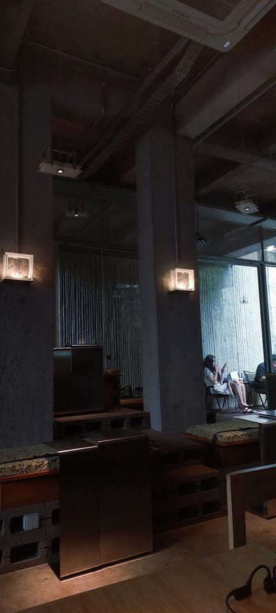 The image depicts the interior of a modern coffee shop, likely in the city of Sampora, Indonesia. The scene is dimly lit, with concrete pillars and exposed ceiling infrastructure suggesting an industrial or loft-style design. Two large square glass block lights are mounted on the concrete pillars, emitting a warm glow. In the background, large windows with vertical blinds allow diffused natural light to enter.

A woman is seated at a table near the window, wearing a white dress and looking down at her phone. Another person is partially visible at a table to her right, wearing a dark shirt. The foreground shows low-profile seating areas constructed from concrete blocks and wooden pallets, topped with patterned cushions. A dark, metallic structure, possibly a counter or table, is positioned in the lower right of the frame. The overall atmosphere is one of quiet contemplation and relaxed social gathering, typical of a contemporary coffee shop environment.
